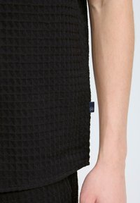 Top negru, texturat, cu un model în grilă. Prezintă o etichetă vizibilă pe latură. Prim-plan al țesăturii cu mâna unei persoane lângă aceasta.
