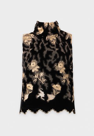 Une femme porte un haut noir sans manches orné d’une broderie florale dorée, un pantalon noir, des escarpins à bout pointu et des boucles d’oreilles créoles, sur un fond clair uni.
