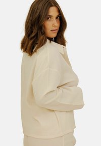 Chemise ample de couleur crème avec un tissu texturé subtil, fermeture boutonnée à l'avant et manches larges, conçue pour le confort et la liberté de mouvement.