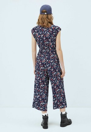 Femme aux cheveux longs portant une combinaison florale bleue, des bottes noires à lacets, des chaussettes blanches et une casquette bleue, se tenant devant un fond blanc uni.