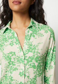 Långärmad skjorta med blommönster i grönt på en krämfärgad bakgrund, med knappstängning fram och strukturerad krage.