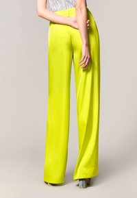 Spodnie z wysokim stanem i szerokimi nogawkami w intensywnym neonowym żółtym kolorze z satyny, charakteryzujące się gładką teksturą i starannie wykończonymi szwami. Tylny widok pokazuje płaski pas.