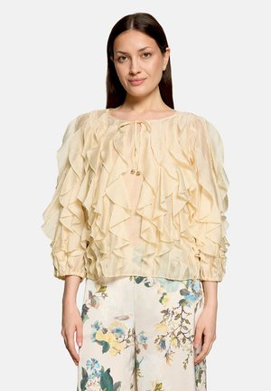 Kvinde med langt mørkt hår iført en lysebeige bluse med flæser og cremefarvede bukser med et blåt og gult blomsterprint, stående mod en ensfarvet baggrund.