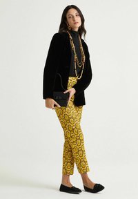 Blazer in velluto nero, dolcevita nero aderente, pantaloni gialli con motivi geometrici, ballerine nere decorate, gioielli con perline, piccola pochette nera.