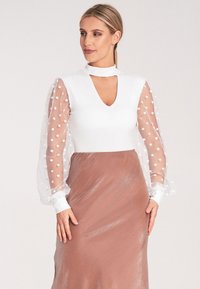 Biała body z dekoltem w serek i długimi przezroczystymi rękawami w kropki, w połączeniu z gładką brązową satynową spódnicą o subtelnej poświacie.