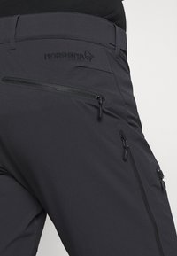 Čierne softshellové nohavice s viacerými zipsovými vreckami, hladkou textúrou a embosovaným logom na pásku.