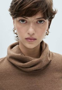 Brun, ribbet turtleneck trøje med løs krave og tekstureret stof. Modellen har kort hår og bærer runde sølvringe. Neutral baggrund.