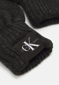 Crne pletene rukavice s teksturiranim materijalom. Imaju crnu oznaku s bijelim "CK" logotipom i natpisom "Calvin Klein Jeans".