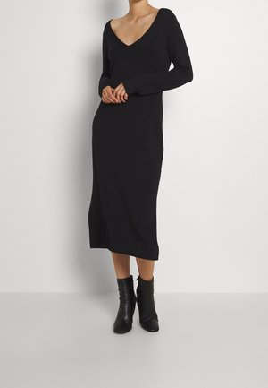 Femme portant une robe midi noire à manches longues avec un décolleté en V profond et des bottines noires, debout devant un fond blanc uni.