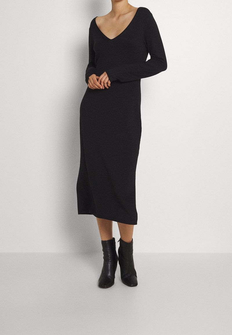Femme portant une robe midi noire à manches longues avec un décolleté en V profond et des bottines noires, debout devant un fond blanc uni.