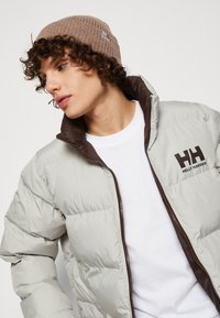 Puffová bunda v světle šedé barvě, se zipem a límcem v hnědé barvě. Logo Helly Hansen je zobrazeno tmavě hnědou barvou na levé straně. Hnědá pletená čepice.