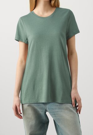 Woman wearing a plain muted green short-sleeve t-shirt and light blue faded jeans, standing against a neutral background.