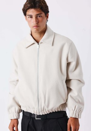 Jeune homme portant une veste blanche zippée avec des poignets et un bas élastiques, debout devant un fond clair uni.