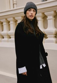 Cappotto di lana nera con collare oversize, berretto in maglia grigia e maglione grigio indossato sotto. Cuffie bianche della camicia visibili sui polsini.