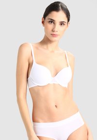 Reggiseno bianco imbottito con spalline sottili e dettagli in pizzo, texture liscia e forma sagomata. Indossato con biancheria intima bianca coordinata.