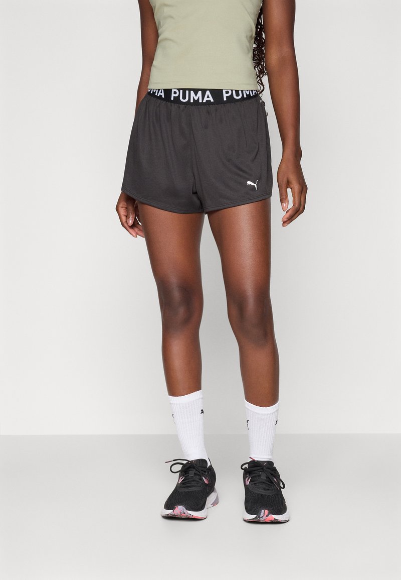 Mustad sportlikud lühikesed püksid, millel on PUMA logo vöö, koos valgete sokkidega ja mustade jooksumudelitega, millel on roosa aktsent.