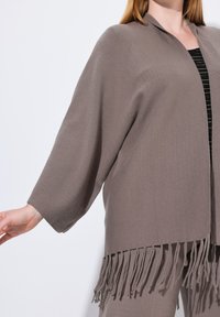 Cárdigan marrón de punto abierto con mangas de 3/4 y detalles de flecos a lo largo del dobladillo. Textura suave y corte relajado.