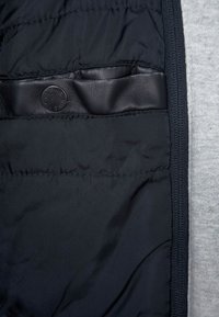 Sort puffjakke med glat foring og læderdetalje. Inkluderer en trykknaplukning mod en grå stofbaggrund.