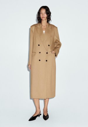 Cappotto beige doppiopetto in misto lana, con grandi bottoni neri, rever a giorno e tasche laterali. Lunghezza al ginocchio con una silhouette dritta.