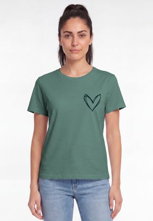 Frau mit dunklen Haaren im Pferdeschwanz, die ein türkisfarbenes T-Shirt mit einem schwarz handgezeichneten Herz auf der linken Brust und hellblaue Jeans trägt.