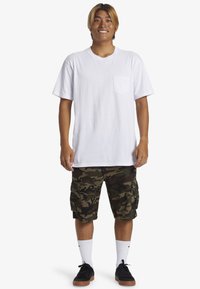Witte T-shirt met korte mouwen en een zak, gecombineerd met camouflage cargo shorts. Draag het met witte crew sokken en zwarte sneakers.