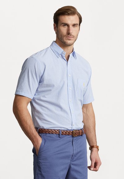 Kortærmet skjorte med knapper i lyseblå med hvide striber, med krave og et logo på brystet, parret med khaki shorts og et flettet bælte.