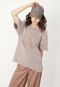 Brązowy, bawełniany t-shirt z efektem prania, o luźnym kroju, z dużym beżowym logo. W połączeniu z dopasowaną brązową czapką z logo.