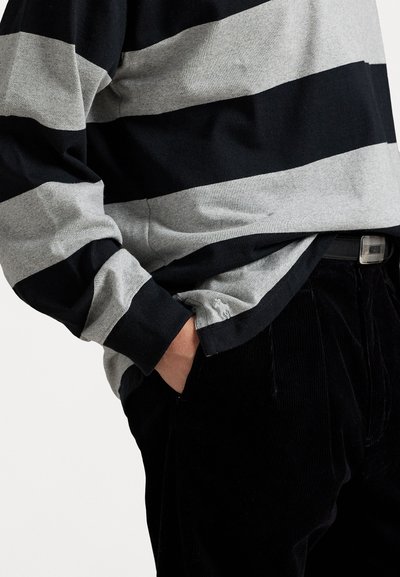 Camisa de manga larga con rayas horizontales en negro y gris, hecha de algodón. Presenta un corte relajado y un bolsillo lateral, combinada con pantalones de pana negros.