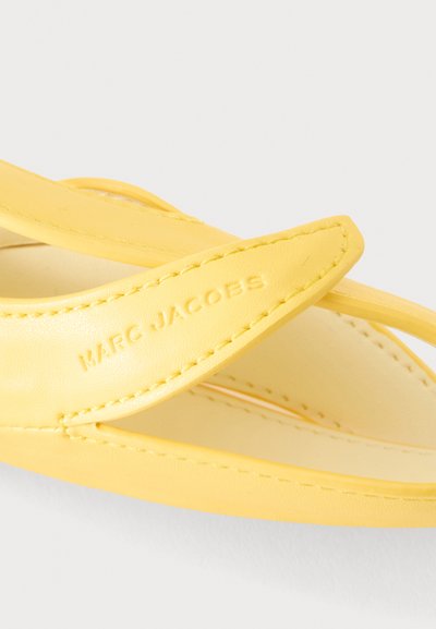 Lanière de sandale en cuir jaune avec bords cousus embossée du logo "MARC JACOBS" en gros plan.