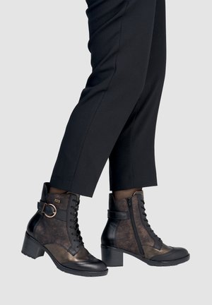 Person wearing black ankle boots with lace-up fronts, side zippers, and buckles, paired with black pants against a plain light background.