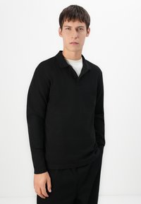 Bărbat tânăr cu părul scurt brunet, îmbrăcat cu un polo negru cu mâneci lungi peste un tricou alb, stând în fața unui fond alb simplu.