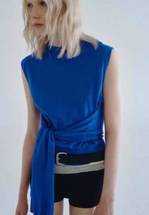 Jeune personne aux cheveux blonds mi-longs portant un haut bleu sans manches noué à la taille et un short noir avec une ceinture noire.