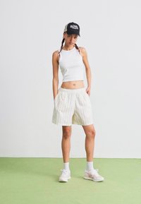 Biely crop top a krémové pruhované šortky, doplnené bielymi teniskami a čiernym šiltovkou. Jednoduchý pozadie s zelenou podlahou.