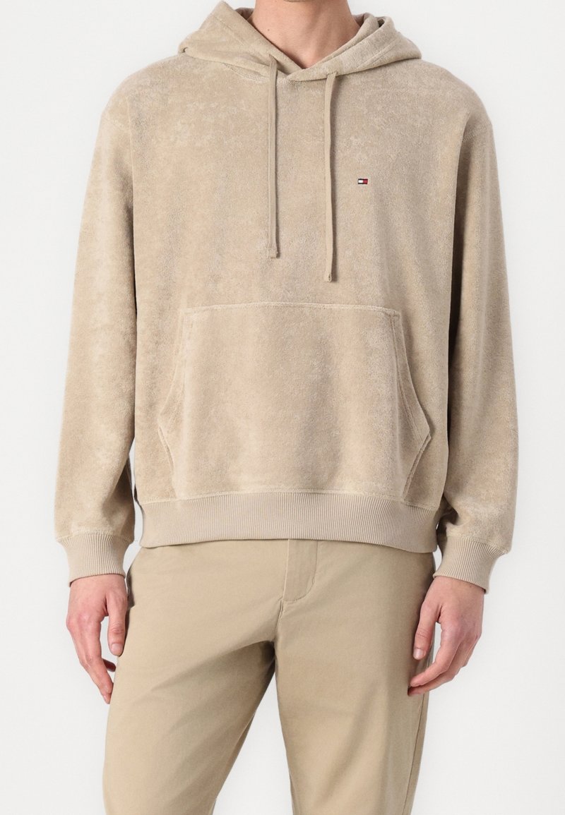 Homme portant un sweat à capuche beige avec poche avant et petit logo sur la poitrine, assorti à un pantalon beige, debout sur fond blanc.