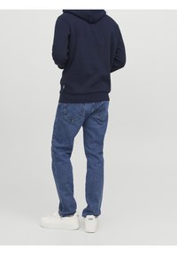 Sudadera marina con bolsillo tipo canguro, vaqueros azules con textura desgastada y zapatillas deportivas blancas con suela texturizada. Costuras y acentos visibles.