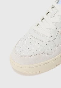 Adidași din piele albă perforată cu accente din ante bej deschis, șireturi plate albe și o talpă din cauciuc texturat.