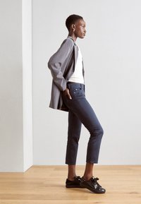 Blazer grigio a maniche lunghe sopra una maglietta bianca, abbinato a pantaloni aderenti blu scuro e sneakers nere con dettagli decorativi e fiocchi.