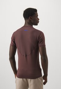 Kortermet, brun treningsskjorte med en tettsittende design. Har meshtekstur på baksiden, blå "VIGOR7"-etikett og glatt, lett stoff.