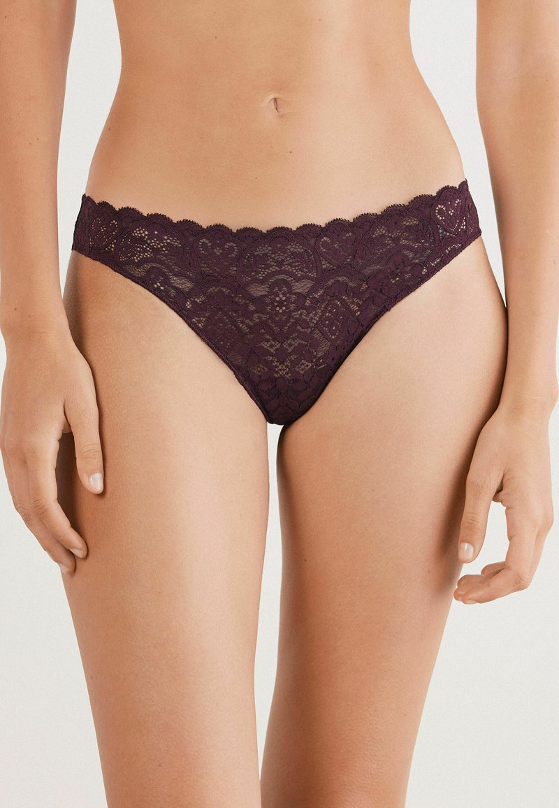 Femme portant un sous-vêtement en dentelle violet foncé, mettant en valeur le milieu du corps et le haut des cuisses contre un arrière-plan clair.