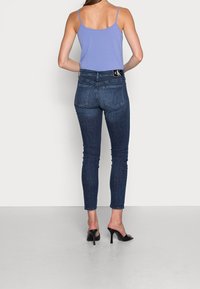 Jeans ajustados de mezclilla azul oscuro con cintura alta y un logo de CK en el bolsillo trasero, combinados con una blusa sin mangas de color púrpura claro y tacones negros.