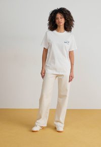 Tricou alb cu mânecă scurtă, având un design grafic mic de culoare albastră pe buzunar, asortat cu pantaloni largi de culoare albă murdară și pantofi albi cu accente portocalii.