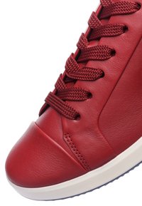 Red leather sneaker with textured surface, featuring braided red laces and a rounded toe, set on a contrasting white rubber sole.