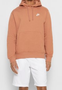 Mikina Nike v teplé terakotově červené barvě s přední kapsou, kapucí na stažení a bílým logem. Nošena s bílými šortkami, nabízí ležérní atletický vzhled.