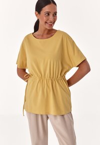 Top de algodón amarillo con un escote amplio, mangas cortas y cintura fruncida. Tela texturizada y detalle de cordón ajustable. Combinado con pantalones claros.