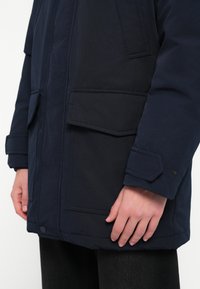 Texturált anyagból készült tengerészkék parkatak, nagy fodros zsebekkel, gombos záródással, valamint állítható mandzsettákkal a nyakrészen és az ujjakon.