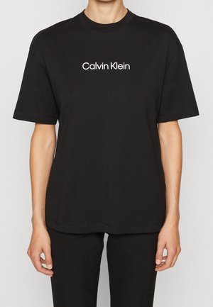 Persona vistiendo una camiseta negra de manga corta con el logo de "Calvin Klein" impreso en blanco en el pecho, combinada con pantalones negros.