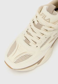 Beige FILA sneakers med netting- og semsket skinnpaneler, tykke kremfargede lisser, og merkevaren sitt logo på tungen mot en hvit bakgrunn.