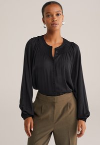 Egy nő fekete, hosszú ujjú blúzt visel, smockolt nyakkivágással és barna, testre szabott, magas derekú nadrággal, egy egyszínű háttér előtt áll.