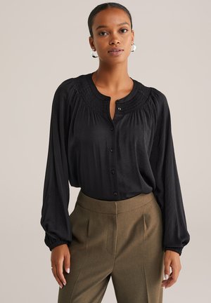 Mujer vistiendo una blusa negra de manga larga con escote fruncido y pantalones marrones de tiro alto, de corte ajustado, de pie contra un fondo liso.