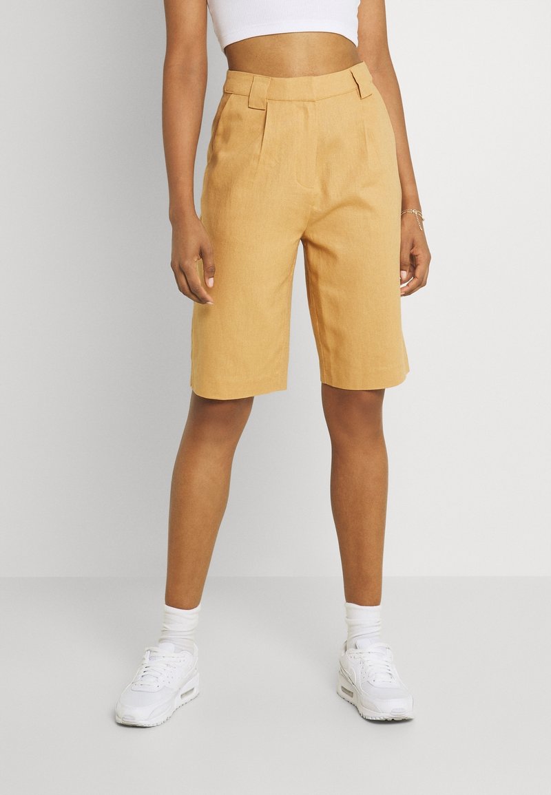 Mustard-colored tailored shorts made of linen blend, featuring pleats and a mid-rise waist. Paired with white sneakers and ankle socks.